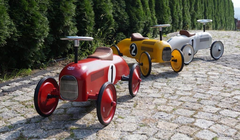Jeździki dla dzieci, w ogrodzie. Na zdjęciu przedstawione są jeździki marki Vilac, metalowe w stylu retro, w trzech kolorach: jeździk czerwony, jeździk żółty, jeździk biały.
